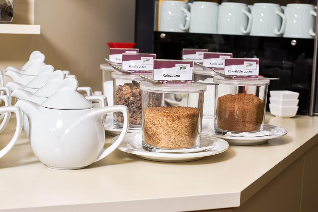 Balta tējas kanna un dažādi cukura veidi stikla traukos uz galda, kā arī porcelāna krūzes.