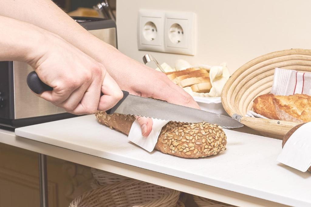 Sagriežot maizi ar asu nazi, redzams rokas darbs virtuvē. Fonā ir dažādas maizes un cepumu izstrādājumi.