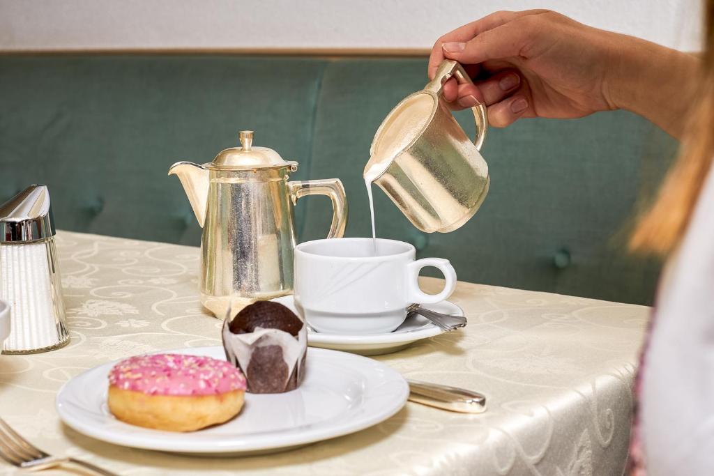 Saldais brokastu galds ar kafijas krūzi, piena kannu un gardiem konditorejas izstrādājumiem, tostarp rožainu donutu un šokolādes kūciņu.