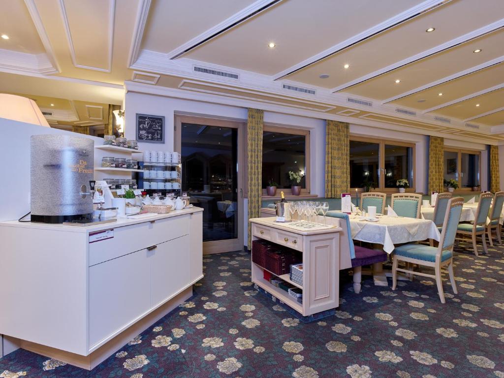 Elegants restorāns ar gaišu interjeru, ko raksturo ziedu raksts uz grīdas un komfortabli galdi ar krēslām. Redzams dzērienu stends un ēdienu pasniegšanas zona ar dažādiem traukiem.