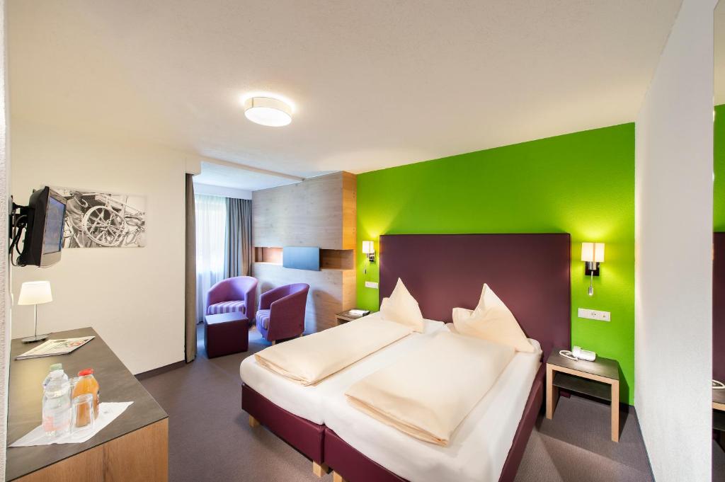 Mūsdienīgs viesnīcas numurs ar spilgti zaļu sienu un ērtām mēbelēm, tostarp divām gultām un atpūtas krēsliem. Telpā ir televizors un galdiņš ar dzērieniem.