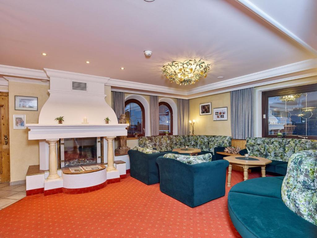 Mājīgs viesistabas interjers ar kamīnu, zaļām dīvānu grupām un siltu apgaismojumu, radot relaksējošu atmosfēru. Sienas rotā bildes, un telpa ir iekārtota ar elegantiem elementiem.