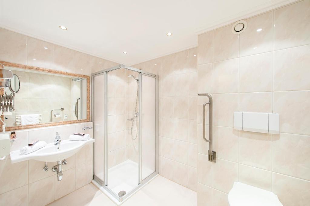 Mūsdienīga vannas istaba ar dušu, izsmalcinātu spoguli un tīru, gaišu flīžu apdari. Vannas istabā ir arī izlietne un higiēnas piederumi.