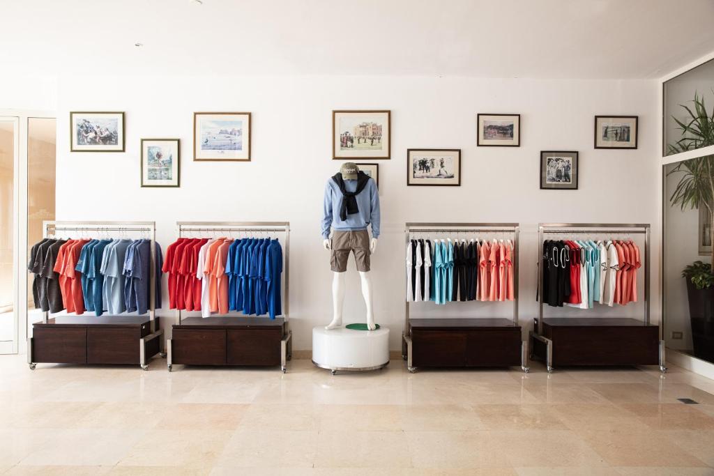 Veikals ar apģērbu stendi, kas piedāvā dažādu krāsu T-kreklus un šortus. Uz pedestāla stāv mannequins, ģērbts stilīgā apģērbā, fona sienā redzami mākslas darbi.