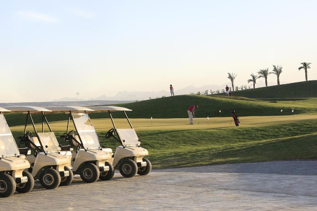 Golfu laukums ar golfa ratiņiem priekšplānā un spēlētājiem fona laukuma zaļumos. Rāmā ainava ar palmām un kalniem tālumā.