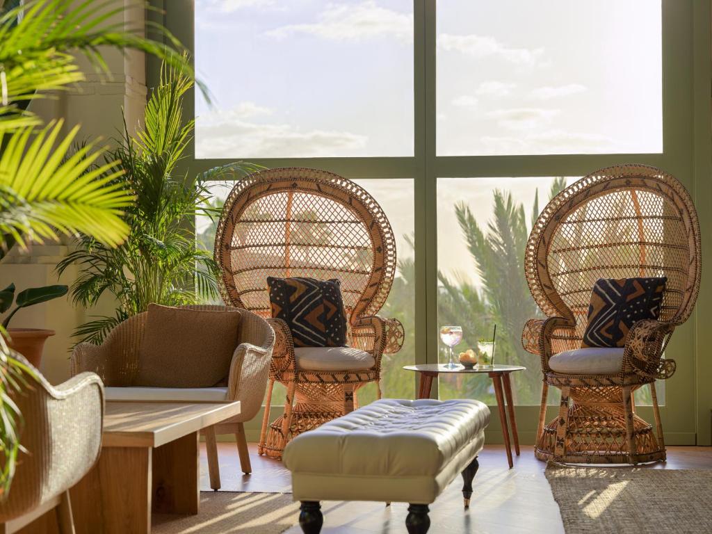 Skaista un gaiša atpūtas telpa ar divām vītņotām krēsliem, zemu galdiņu un ērtu dīvānu, kas apņem zaļas palmas. Gaisma plūst caur lielajiem logiem, radot relaksējošu atmosfēru.