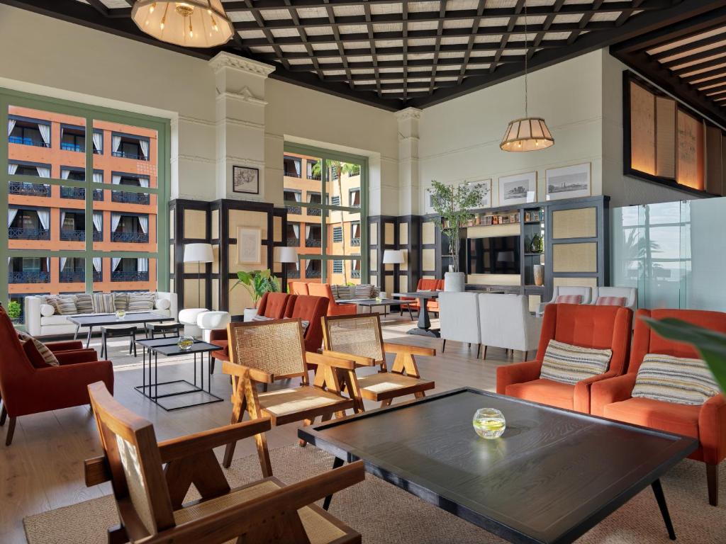 Mūsdienīgs viesnīcas viesistabas interjers ar sarkaniem dīvāniem, koka krēsliem un lieliem logiem, kas nodrošina dabisko apgaismojumu. Viegla un eleganta atmosfēra ar augu akcentiem un mūsdienīgu mēbeļu dizainu.