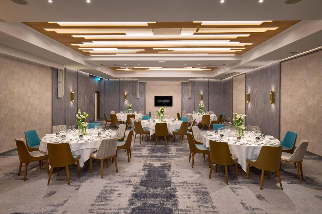 Labi iekārtota banketu zāle ar elegantiem galdiem, ziediem un mūsdienīgu apgaismojumu, ideāli piemērota pasākumiem un svinībām.