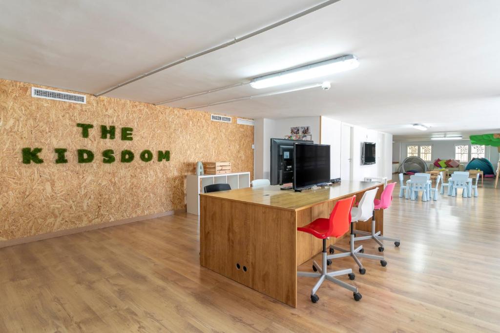 Bērnu aktivitāšu telpa ar nosaukumu "THE KIDSDOM", kas iekārtota ar krāsainiem krēsliem un galdu, nodrošinot radošu vidi. Ir redzami bērnu galdi un telpas atpūtas zonas.