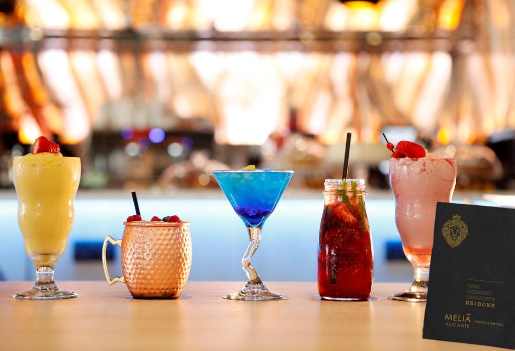 Krāsaini kokteiļi, tostarp augļu un zilo dzērienu variācijas, izvietoti uz bāra, blakus Melia Alicante viesnīcas brošūrai.