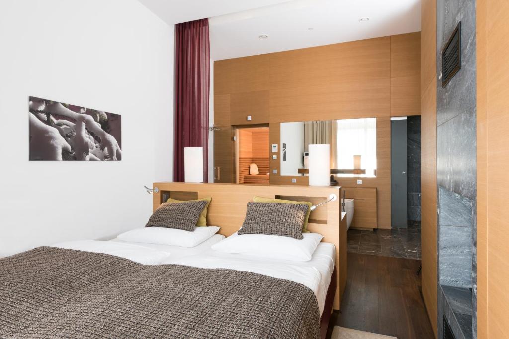 Mūsdienīga guļamistaba ar divām gultām, koka apdari un elegantu interjeru, kas ietver lielu spoguli un modernus apgaismojuma risinājumus. Guļamistabā ir arī mākslas darbs pie sienas un logi ar aizkariem.