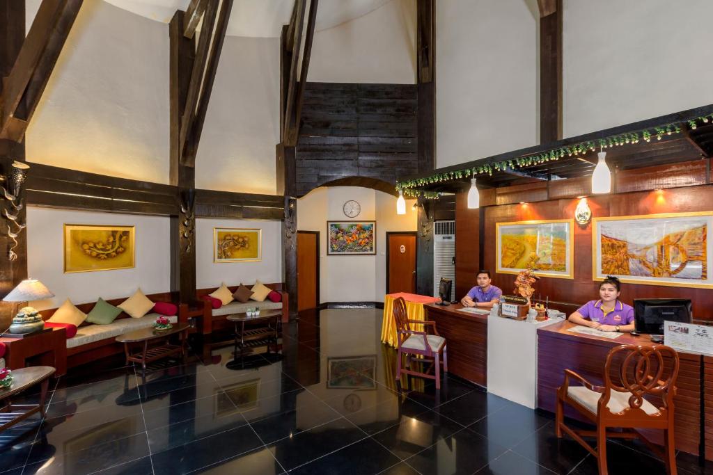 Mūsdienīgs viesnīcas reģistratūras interjers ar augstiem jumtiem, koka konstrukcijām un siltu apgaismojumu, kur darbinieki sagaida viesus. Eleganti iekārtots gaidīšanas zona ar dīvāniem un mākslas darbiem uz sienām.