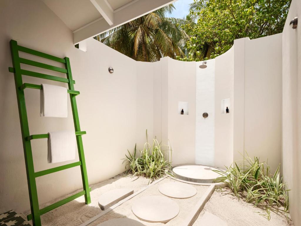 Āra duša ar zaļu dvieļu turētāju un gaišām sienām, apkārt aug zāle un palmas, radot relaksējošu atmosfēru.