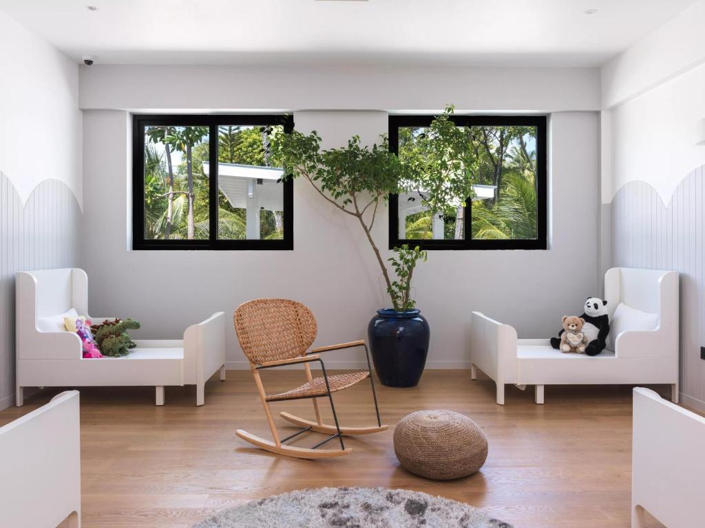 Mūsdienīgs bērnu istabas interjers ar baltām mēbelēm, koka grīdu un augiem. Telpā ir divas gultas, šūpuļa krēsls un dekoratīvi rotaļlietu elementi.