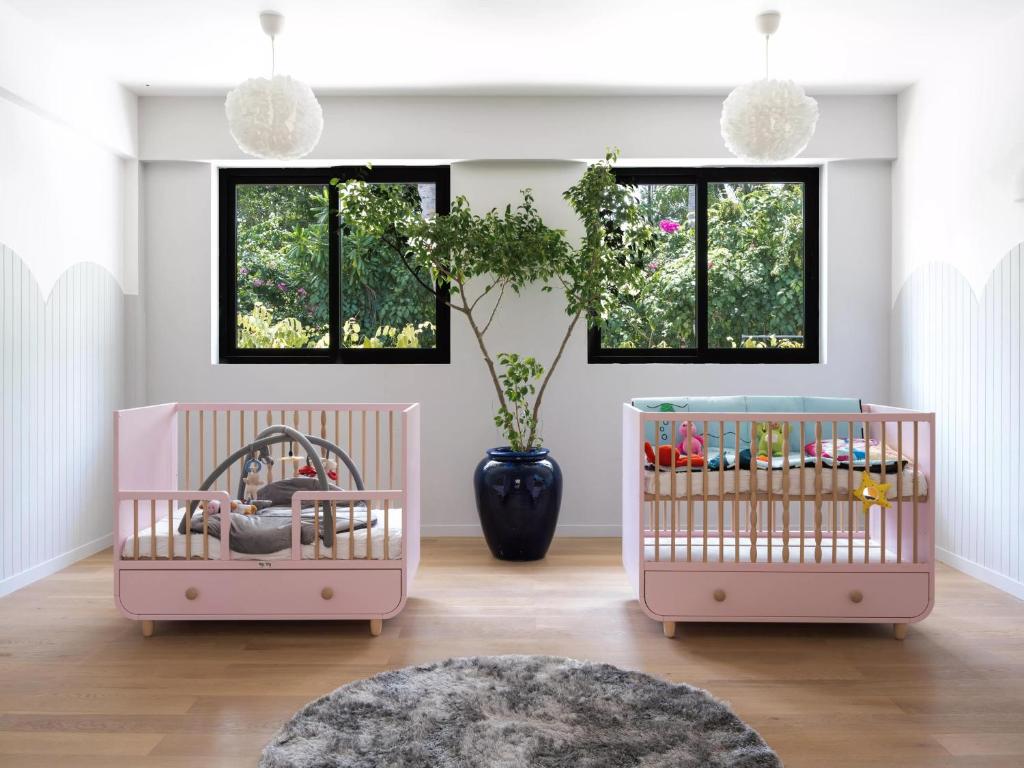 Mūsdienīgs bērnistabas interjers ar divām rozā gultiņām un zaļo augu centrā. Labi apgaismots telpas ar lieliem logiem un koka grīdu.