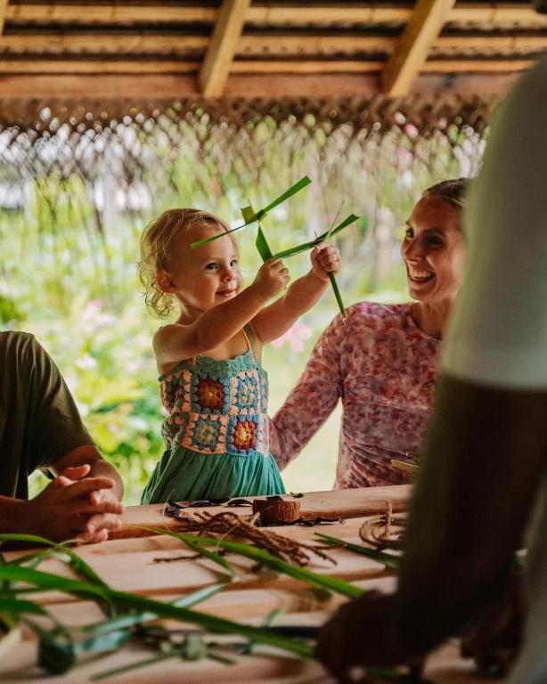 Bērns ar prieku spēlējas, turot zaļās lapas, kamēr pie galda sēž pieaugušie, kas smaida un vēro viņu radošās aktivitātes. Apkārt ir zaļa daba un radoša atmosfēra.