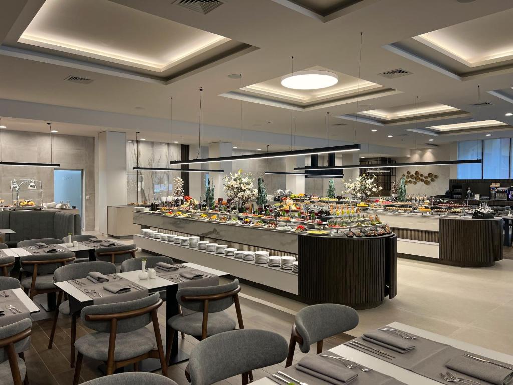 Mūsdienīgs restorāns ar plašu bufeti, kurā redzami dažādi ēdieni un saldumi, eleganti galdi un krēsli. Apgaismojums rada patīkamu atmosfēru.
