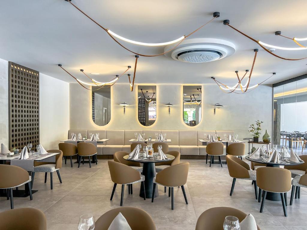 Moderns restorāns ar elegantu interjeru, kas iekārtots ar stilīgām mēbelēm un gaismas ķermeņiem. Telpā redzami galdi ar salvetēm un traukiem, radot patīkamu atmosfēru.