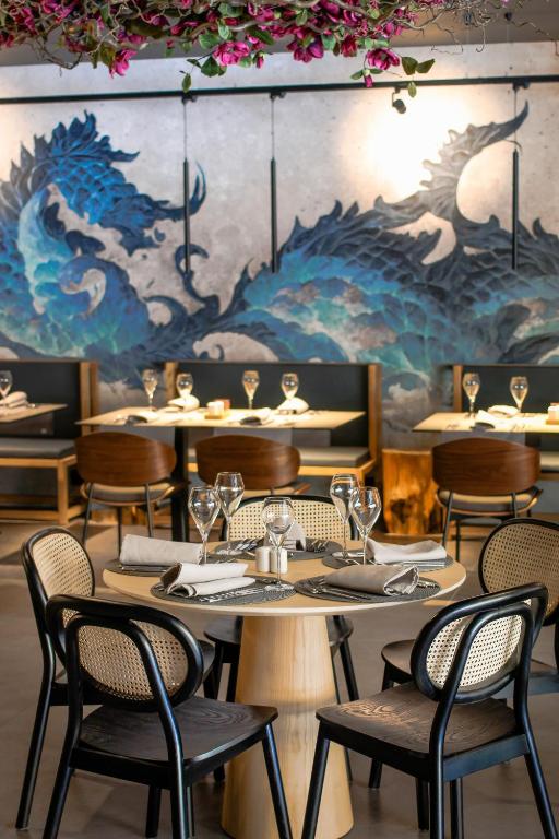 Mūsdienīgs restorāns ar elegantu interjeru, centrā redzams apaļs galds ar krēsliem un dekoratīvu sienas gleznojumu, kas attēlo viļņus.