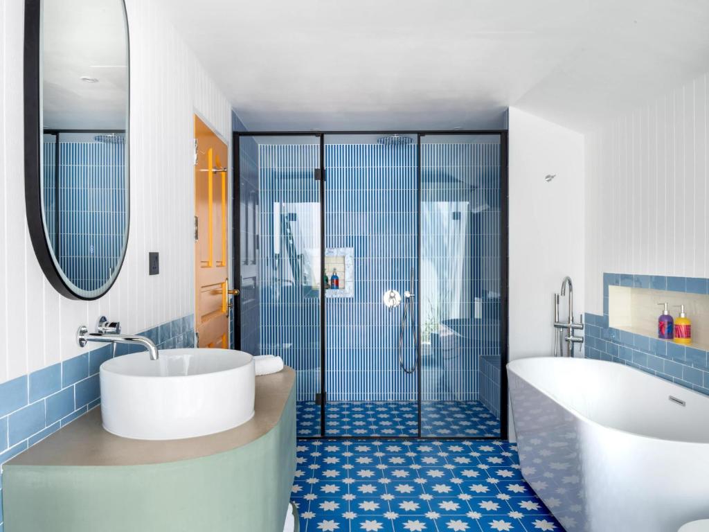 Mūsdienīgs vannas istabas interjers ar zilām flīzēm, stilīgu vannu un modernas dušas kabīni. Telpa ir gaiša un eleganta, ar ovālu spoguli un krāsainiem higiēnas līdzekļiem.