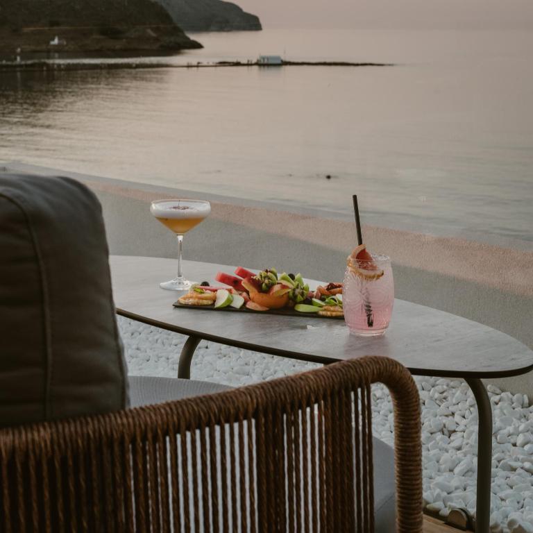 Rāmja ar skatu uz mierīgu ūdeni, kur uz galda ir dažādi augļi un dzērieni, radot relaksējošu atmosfēru. Ideāla vieta atpūtai pie jūras.