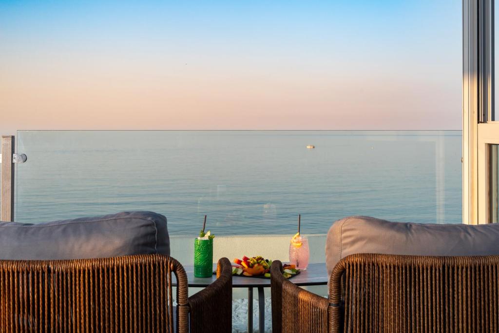 Skats no balkona uz mierīgu jūru, kur uz galda atrodas divi kokteiļi un augļu šķīvis, radot relaksējošu atmosfēru.