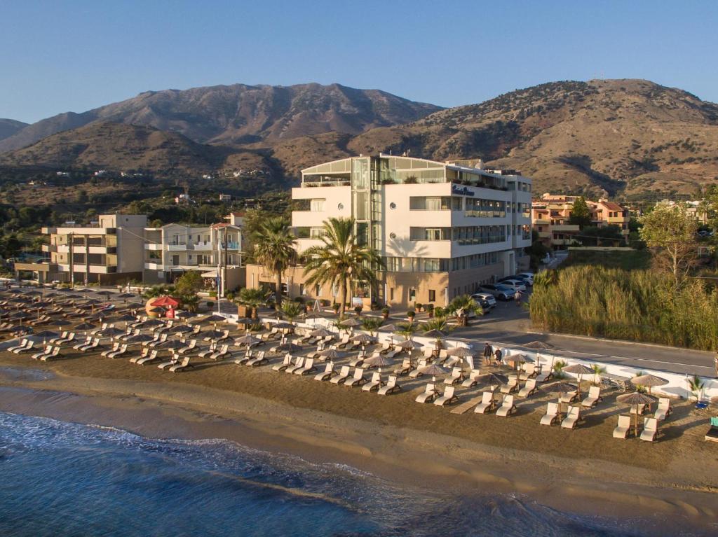 Pludmales viesnīca ar modernu arhitektūru, kas atrodas pie ūdens, ap kuru ir izvietoti sauļošanās krēsli un palmas, un fona veido kalni.