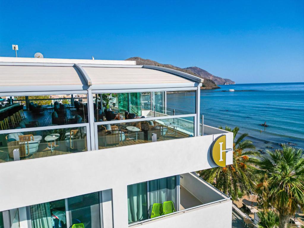 Mūsdienīga viesnīcas terasa ar skatu uz jūru, kurā redzami brīvi sēdoši apmeklētāji un palmām apstādīta piekrastes ainava.