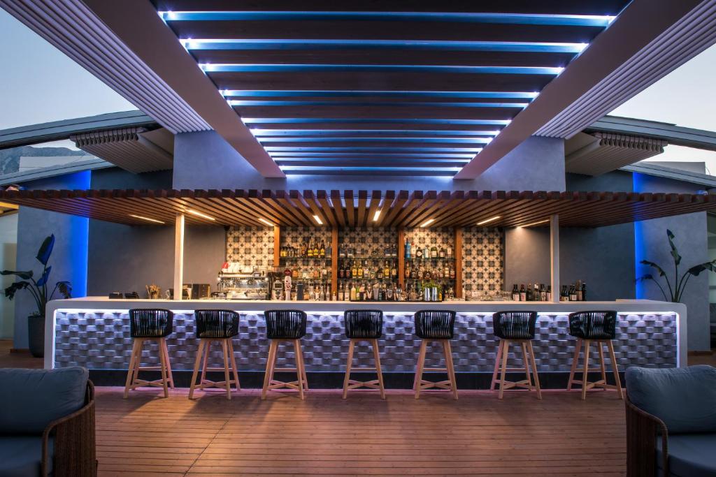 Moderns bārs ar elegantiem krēsliem un akcentējošu apgaismojumu. Aiz bāra redzama plaša alkohola izvēle un stilīgs interjers.