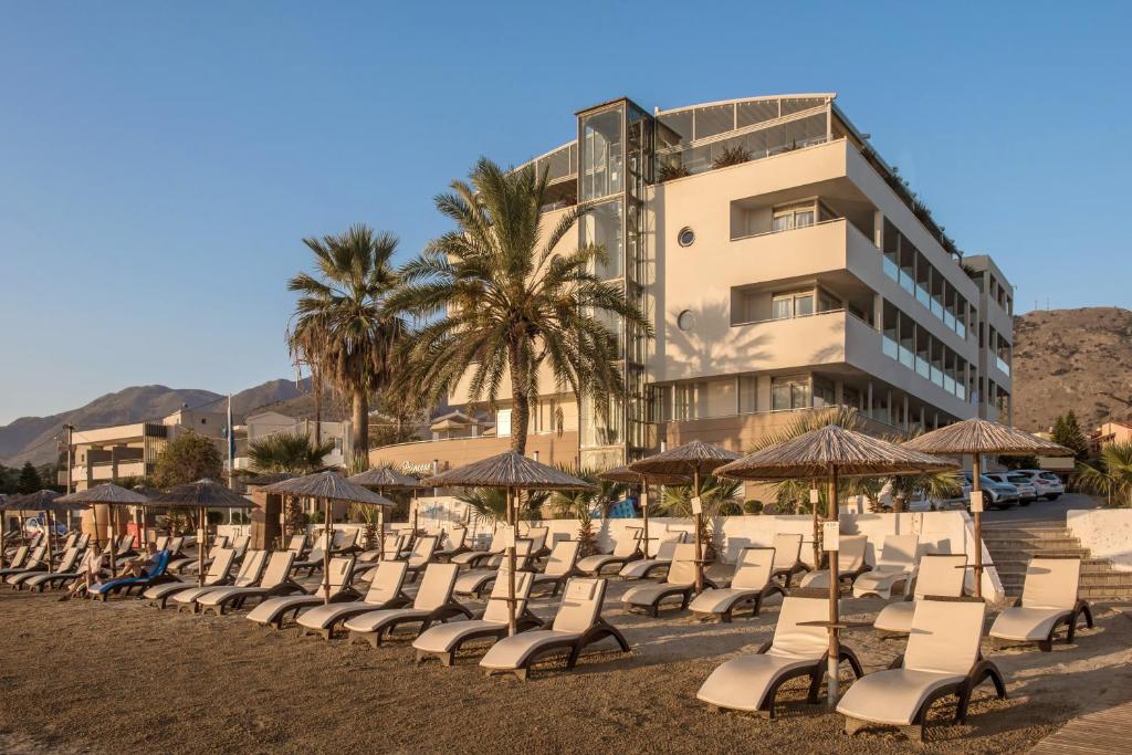 Pludmales skats ar saulē apsildītu viesnīcu fonā, kur atrodas pludmales krēsli un palmas. Ideāla vieta atpūtai un relaksācijai.