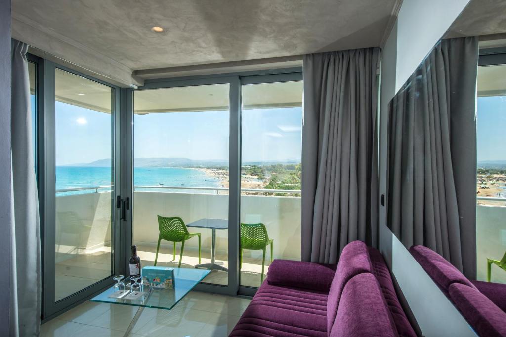 Mūsdienīga viesnīcas istaba ar skatu uz jūru, kurā ir purpursarkans dīvāns, stikla galds un zaļas krēsli uz balkona. Gaismas un plūstošs interjers rada relaksējošu atmosfēru.