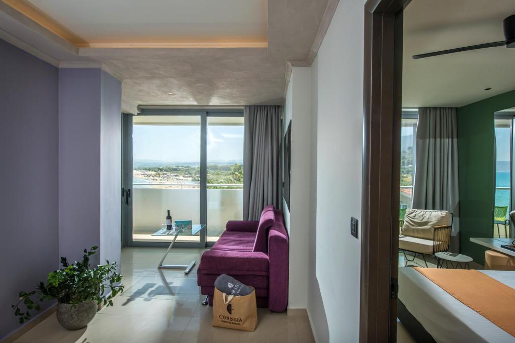 Mūsdienīgs viesnīcas numurs ar skatu uz jūru, kurā ir purpura dīvāns un stikla galdiņš. Numurs ir gaišs un elegants, ar zaļām un neitrālām krāsām.