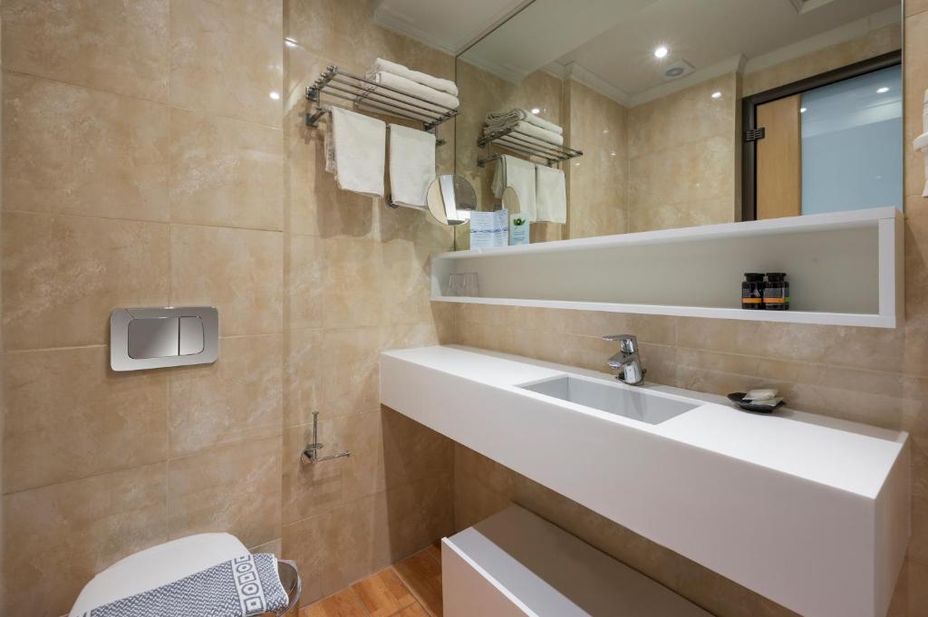 Mūsdienīgs vannas istabas interjers ar gaišām flīzēm, elegantu izlietni un uzglabāšanas plauktu. Vannas istabā ir arī dvieļi un higiēnas piederumi.