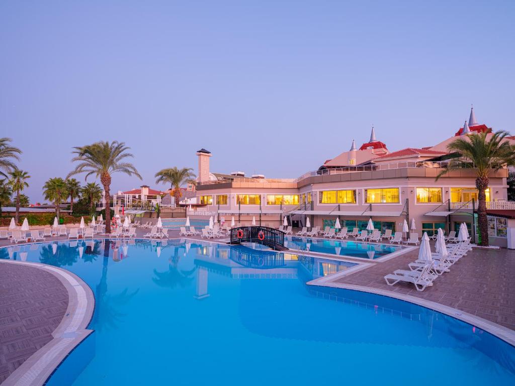 Aydinbey Famous Resort baseins ar palmām un ēku pie saulrieta.