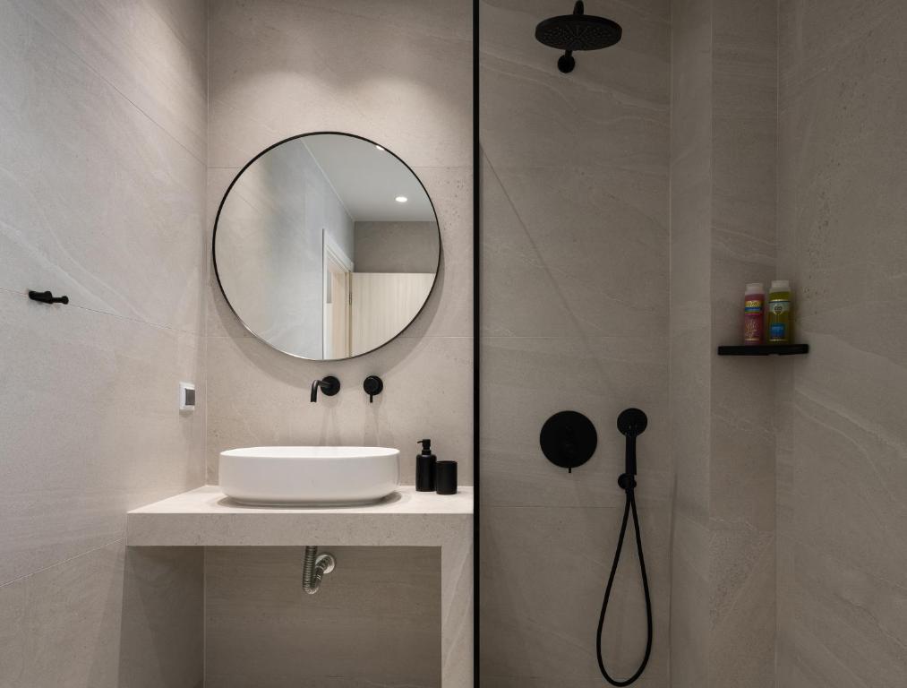 Mūsdienīgs vannas istabas interjers ar apaļu spoguli, minimālistisku izlietni un melnu dušas ierīci uz gaišas akmens sienas.