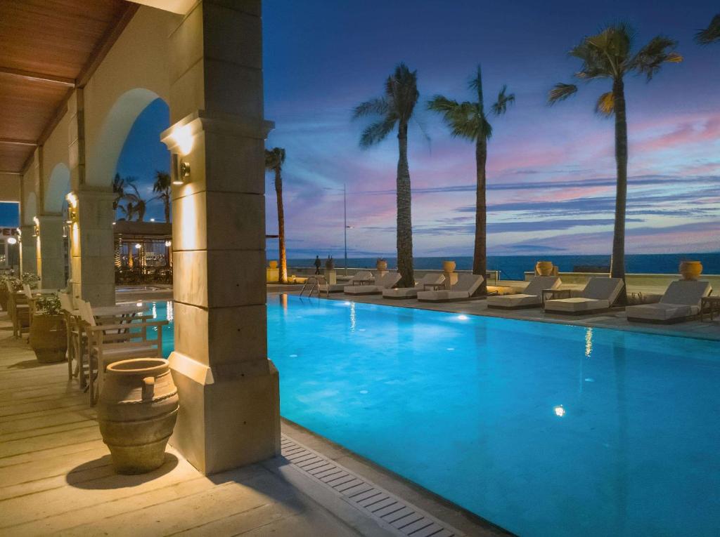 Grecotel Plaza Beach House baseins ar palmām un saulrietu piekrastē.