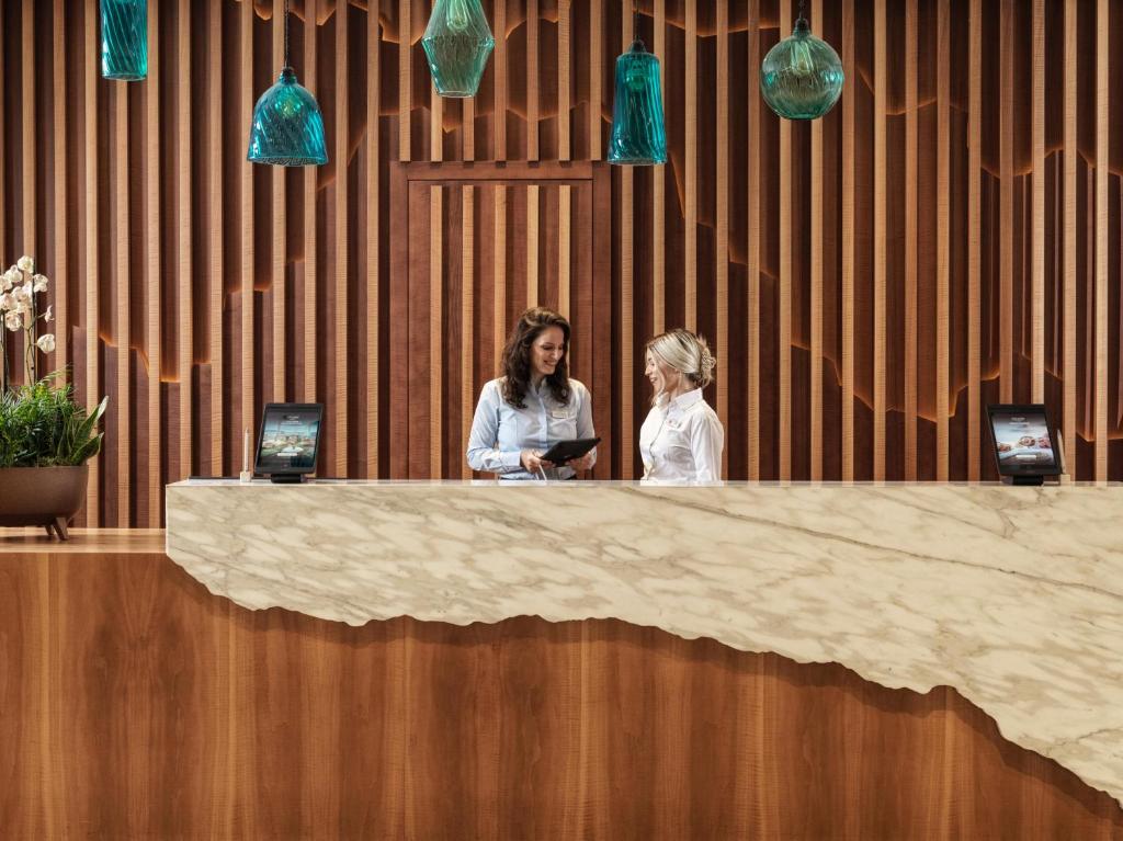 Mūsdienīgs viesnīcas reģistratūras interjers ar koka akcentiem un stilīgām gaismas ķermeņiem, kur divas darbinieces sarunājas pie marmora reģistratūras.