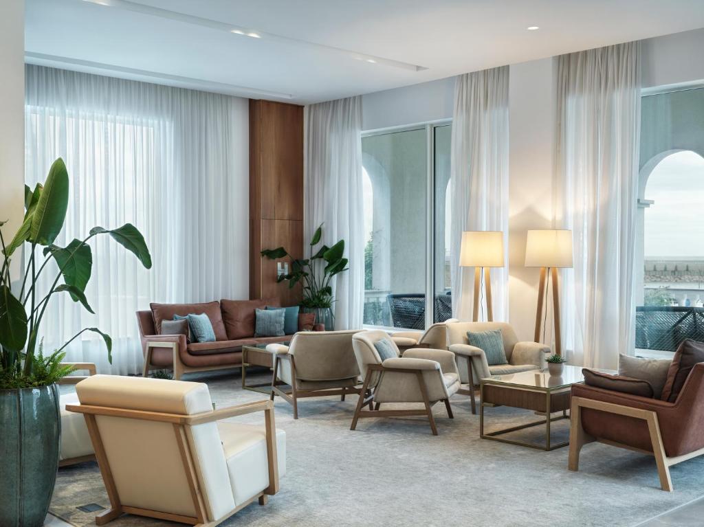 Moderns viesistabas interjers ar komfortablām mēbelēm, augiem un lieliem logiem, kas nodrošina dabisko apgaismojumu.