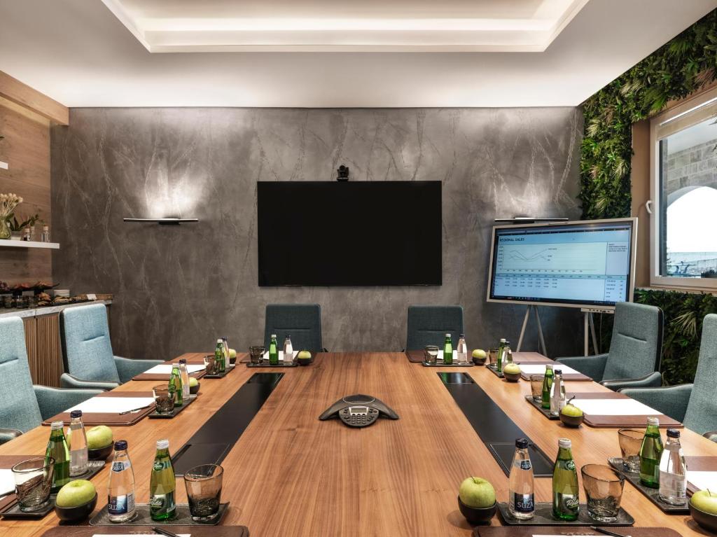 Mūsdienīga sanāksmju telpa ar koka galdu, zilo krēslu rindām un tehnoloģiju, tostarp ekrānu un videokonferenču iekārtu. Telpā ir pieejami dzērieni un veselīgas uzkodas.