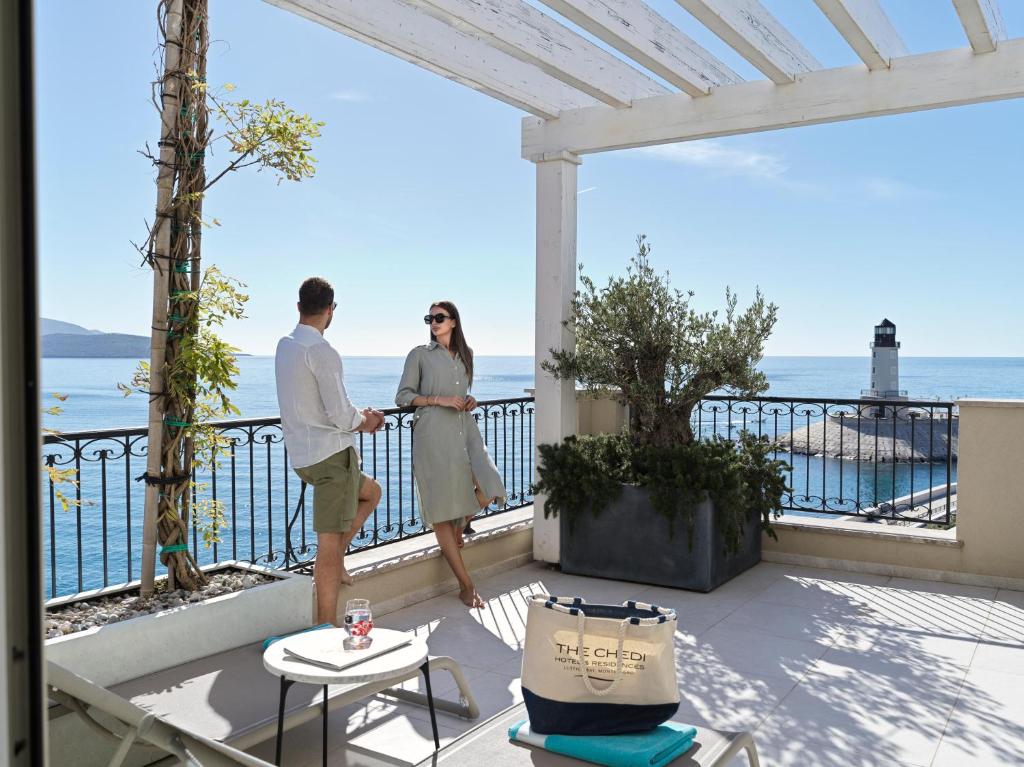 Divi cilvēki atpūšas terasē ar skatu uz jūru un bāku, baudot saulaino dienu. Terasē ir zaļumi un mājīgas mēbeles, radot relaksējošu atmosfēru.