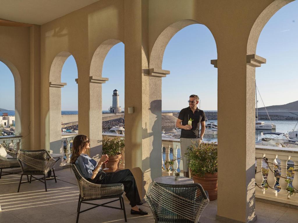 Divi cilvēki atpūšas terasē ar skatu uz ostu, kur redzami jahtas un bāka. Gaisma ir silta un radoša atmosfēra veido mierīgu brīvdienu noskaņu.