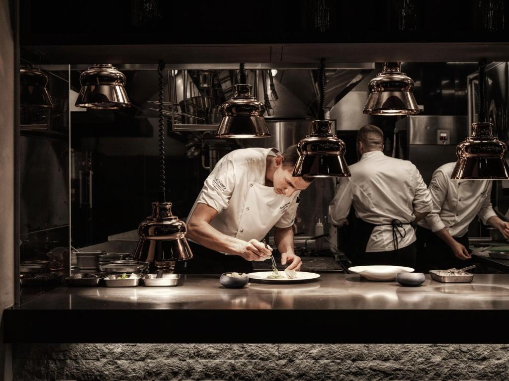 Šefpavārs gatavo ēdienu modernā restorānā, izmantojot elegantas virtuves iekārtas un apgaismojumu. Fons rāda citus pavārus, kas strādā kopā.