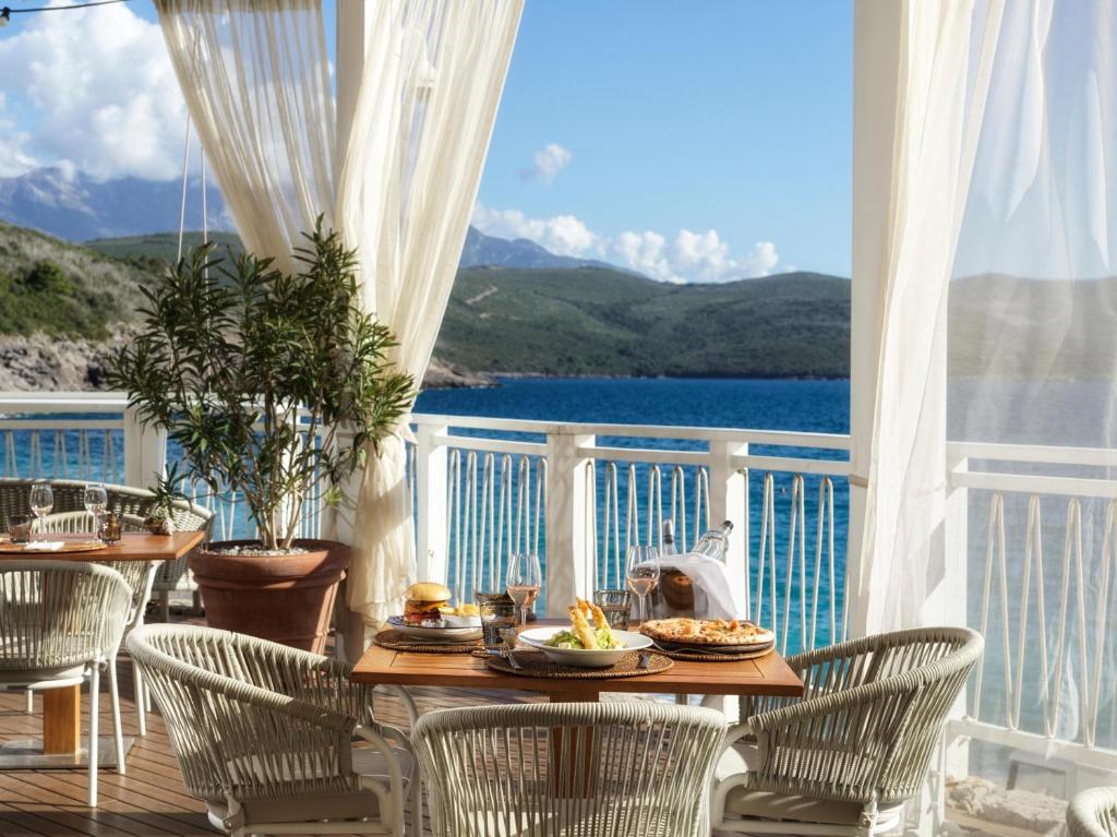 Jūras piekrastes restorāna terase ar galdiem, kas klāti ar ēdieniem un dzērieniem, un skatu uz zilo ūdeni un kalniem. Viegli aizkaru plīvuri piešķir vietai romantisku atmosfēru.