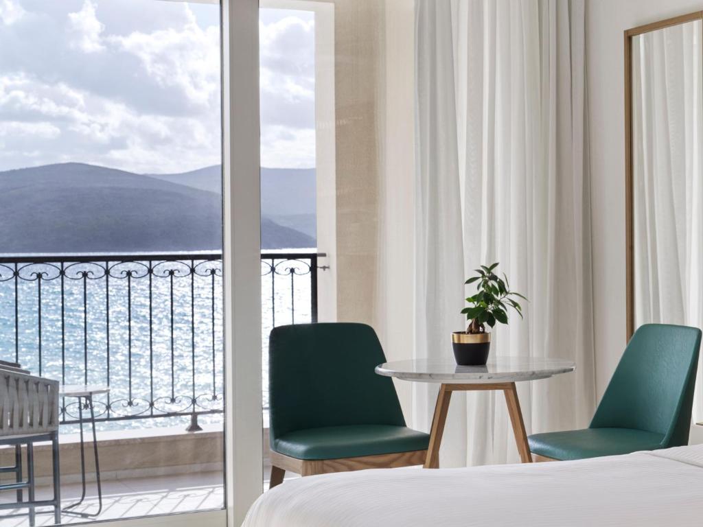 Mūsdienīgs viesnīcas numurs ar skatu uz jūru, kurā ir divi zaļi krēsli, apaļš galds un neliela auga. Gaismas plūst caur lielajiem logiem, radot relaksējošu atmosfēru.