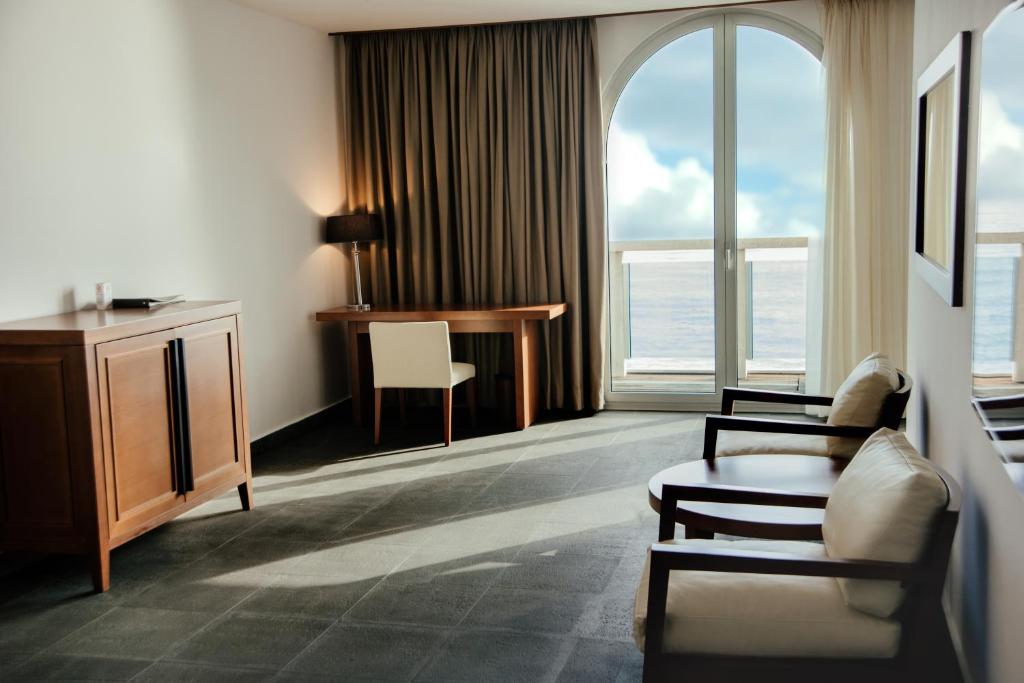Mūsdienīgs viesnīcas numurs ar skatu uz jūru, kurā ir koka mēbeles, rakstāmgalds un komfortabli krēsli. Labi apgaismots ar lieliem logiem un aizkariem.