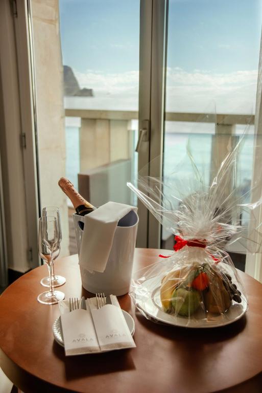 Galda dekorācija ar šampanieti, glāzēm un augļu grozu, kas izlikta blakus logam, no kura paveras skats uz jūru.