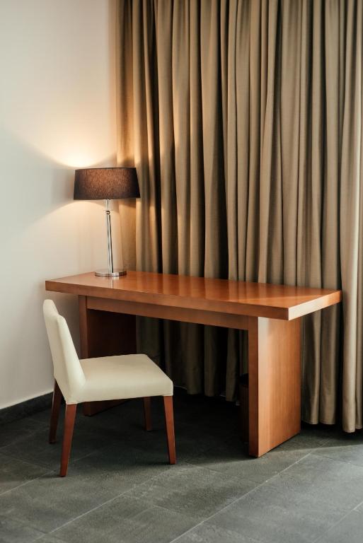 Koka galds ar modernu lampu un krēslu, blakus ir drapētas aizkaru. Telpa ir gaiša un eleganta.