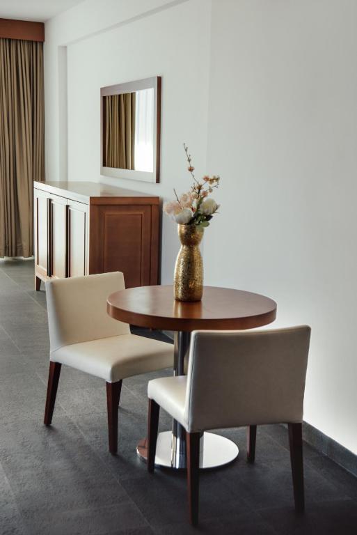 Mūsdienīgs interjera dizains ar apaļu galdu un divām krēslām, dekorēts ar ziediem vāzē. Fons ietver aizkarus un spoguli, radot elegantu atmosfēru.