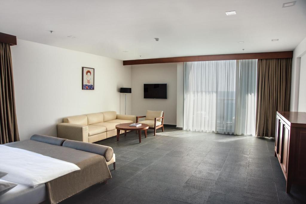 Gaišs un moderns viesnīcas numurs ar dīvānu, galdu un televizoru. Lielas logu žalūzijas ļauj ienākt dabiskajai gaismai.