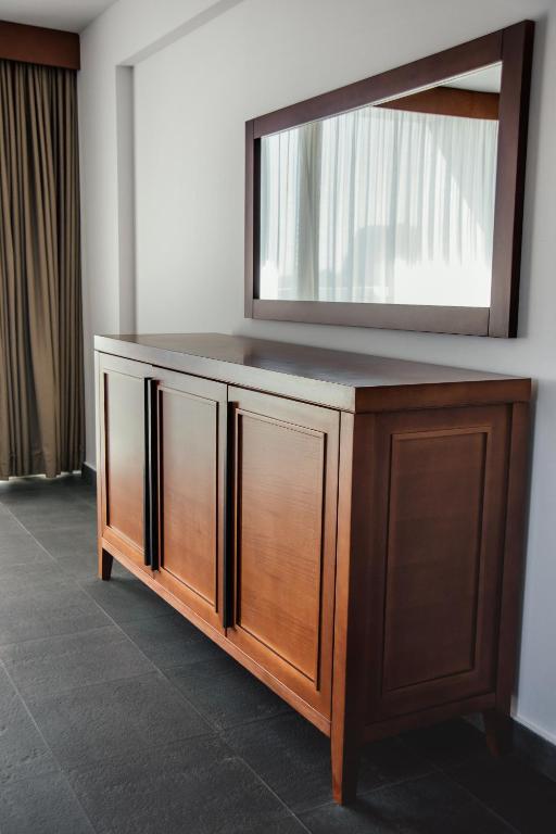 Koka kumode ar divām durvīm un spoguli uz sienas, kas atrodas telpā ar gaišiem aizkariem un melnu grīdu.